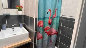 A bathroom with a white sink, dark tiles, a mirror, and a shower curtain with flamingos. - Apartment 46 (Agde)