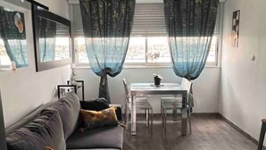 A modern living room with a grey sofa, a dining table with chairs, sheer blue curtains, and a patterned rug. - Apartment 46 (Agde)