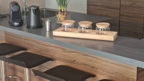 Una cocina moderna con un hervidor de acero inoxidable, una cafetera y una planta en maceta sobre la encimera. Hay cuatro frascos de vidrio con tapas de madera sobre una bandeja de madera.