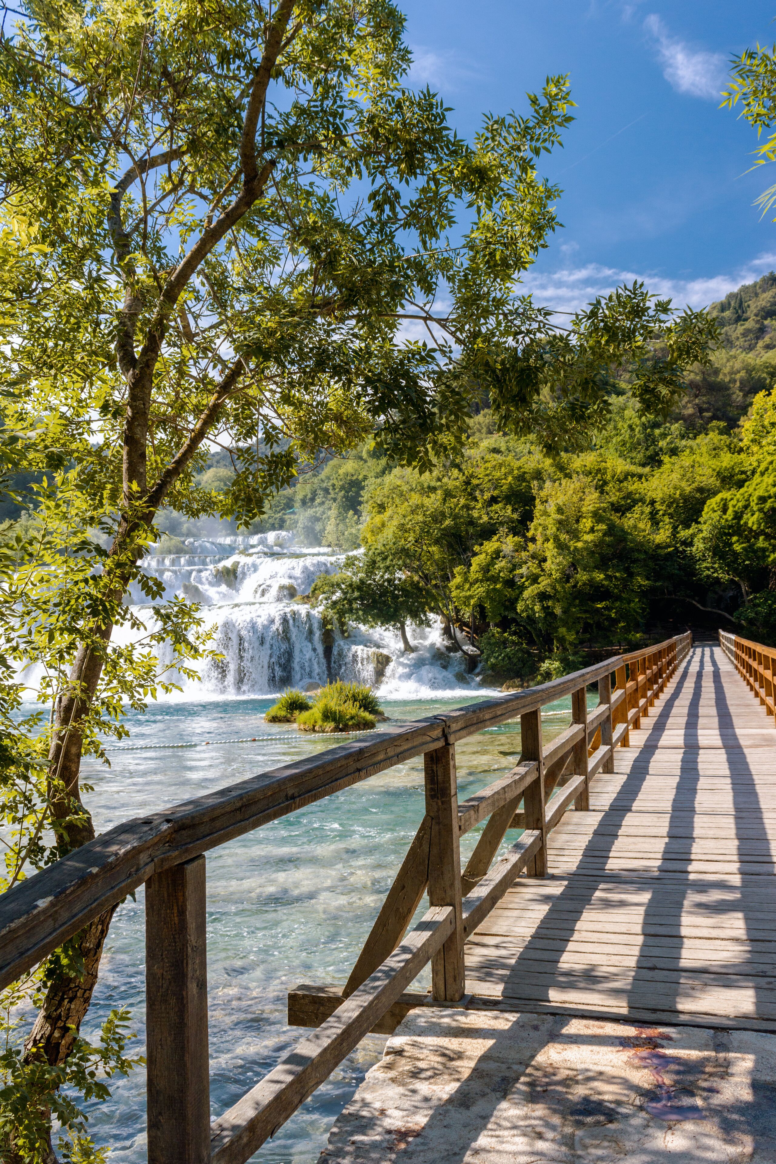 A wooden bridge with a railing leading to a waterfall surrounded by lush greenery.