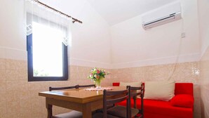 Un coin repas avec une table et des chaises en bois, un canapé rouge, une fenêtre ornée de rideaux en dentelle et un appareil de climatisation.