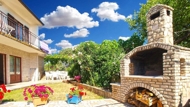 A stone-built outdoor kitchen with a chimney, surrounded by a garden with flowering plants and a paved patio. - Holiday apartment near the beach (Pješćana Uvala)