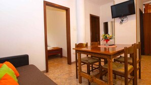 A hotel room with a wooden dining table, four chairs, a sofa with colorful cushions, a TV mounted on the wall, and a flower vase on the table.