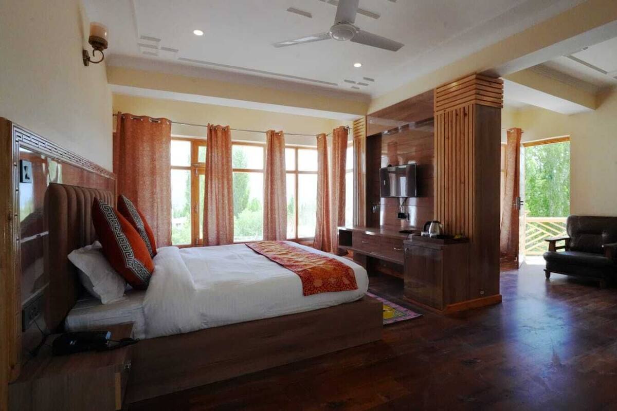 A spacious bedroom with a large bed, wooden furniture, a TV, and a balcony with a chair.
