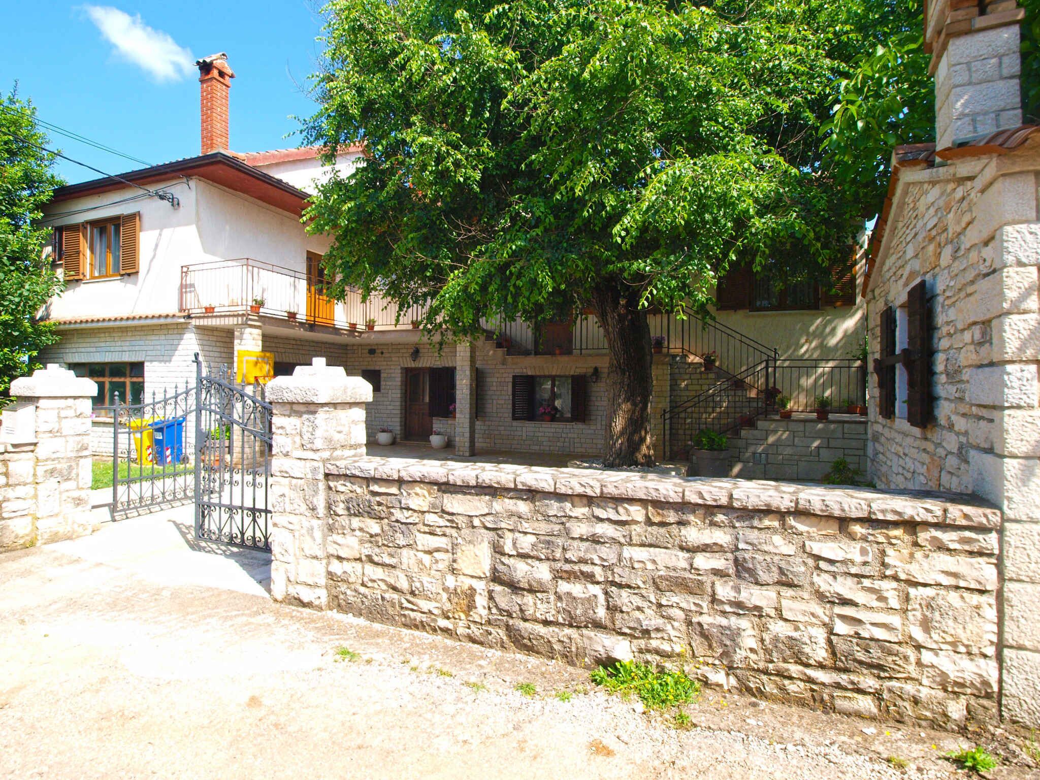 A stone-walled building with a large tree in front, a metal gate, and a staircase leading to a second floor.