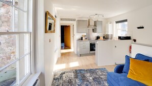 A modern kitchen with grey cabinets, a built-in oven, and a microwave. There is a blue sofa with a yellow pillow in the living area. - Little Edge: Bright and Modern Central Apartment (Berwick-upon-Tweed)