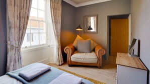 A room with a bed, a chair with cushions, a desk with a laptop, a lamp, and a window with curtains. - Little Edge: Bright and Modern Central Apartment (Berwick-upon-Tweed)