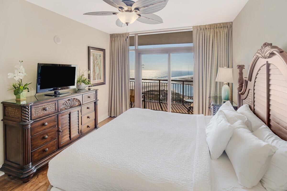 Amplia habitación de hotel con una cama grande, un televisor sobre una cómoda de madera, un balcón con vistas y un ventilador de techo.