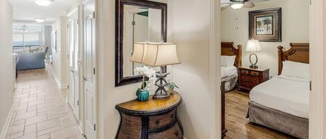 Un couloir d’hôtel avec un plancher en bois, un miroir, une table de chevet avec une lampe et un lit avec une tête de lit.