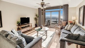 Ein Wohnzimmer mit zwei grauen Sofas, einem Glastisch, einem Fernseher und einem Balkon mit Meerblick.