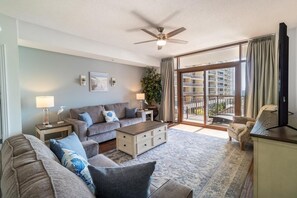A spacious living room with a sofa set, a coffee table, and a television. There is a ceiling fan and a large window with a view of the city. - Seaside Serenity In a 2 Bedroom Condo Amazing View (North Myrtle Beach)