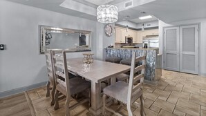 A dining area with a wooden table and chairs, a chandelier, and a kitchen in the background.