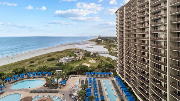 A high-rise hotel with multiple balconies overlooking a beach and a pool area with sun loungers and umbrellas.