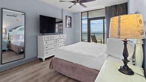 A hotel room with a bed, a dresser, a flat-screen TV, a ceiling fan, a large window with a view, and a lamp with a decorative shade.