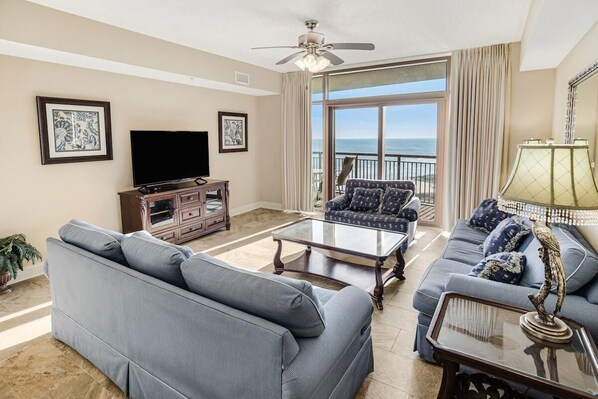 A living room with a large sofa, a coffee table, a TV, and a balcony with a view of the ocean.