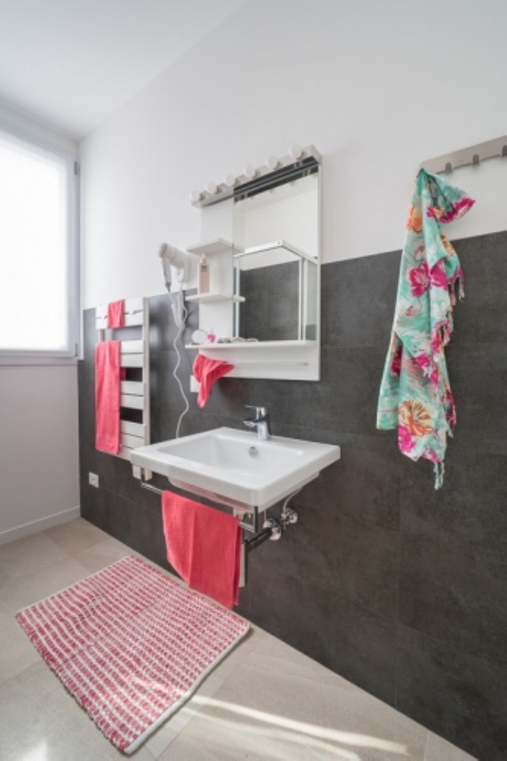 A modern bathroom with a white sink, dark tiled walls, and a red towel on the floor.
