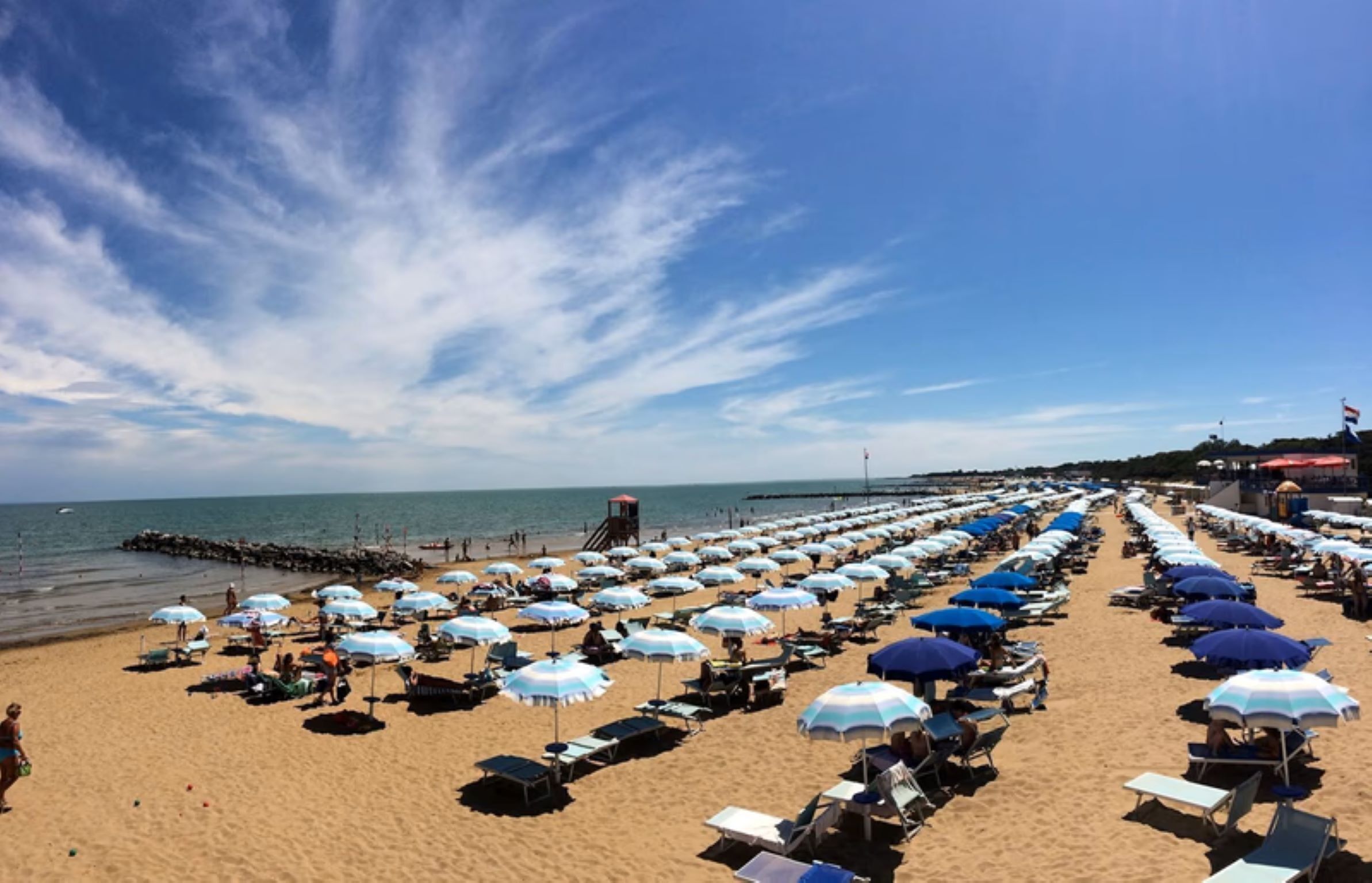 Una playa con hileras de sombrillas y tumbonas, un muelle que se adentra en el agua, y un cielo azul despejado.
