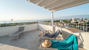 A balcony with two lounge chairs, one with a hat and a blanket, and a view of the sea and cityscape.