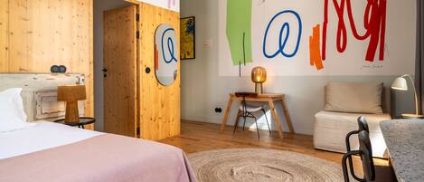 A modern hotel room with a wooden floor, a bed with a pink bedspread, a round rug, a small wooden table with a chair, and a wall with abstract art.