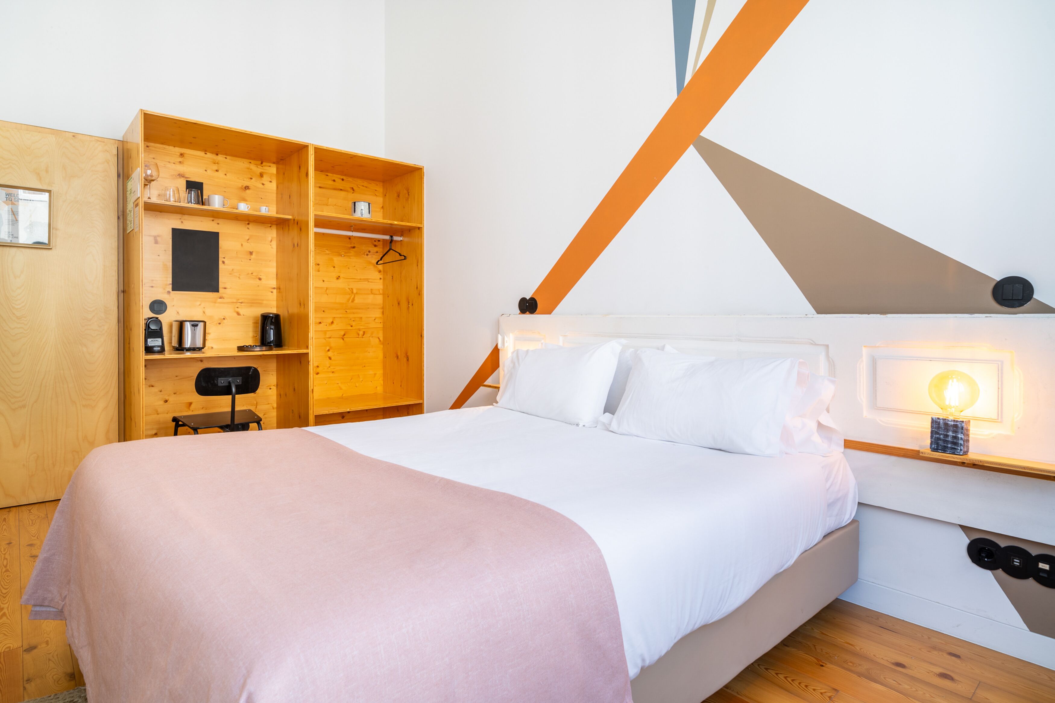 A bedroom with a bed, a wooden wardrobe, a desk with a chair, and a wall-mounted light.