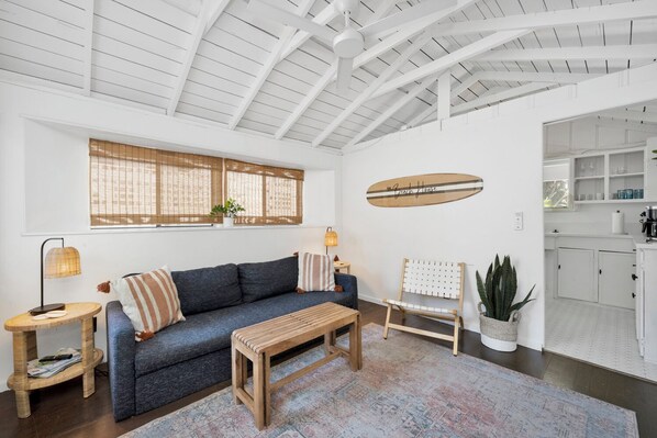 A living room with a sofa, a wooden coffee table, a chair, a window with blinds, a wall-mounted surfboard, and a kitchen visible through an open door.