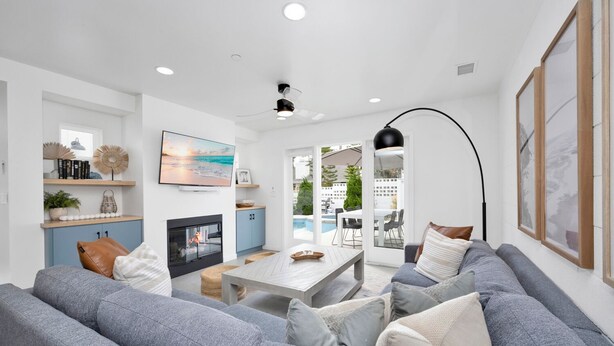 Ein modernes Wohnzimmer mit grauen Sofas, einem Holztisch und einem Fernseher an der Wand. Es gibt einen Kamin, einen Essbereich und einen Blick auf einen Pool im Freien.