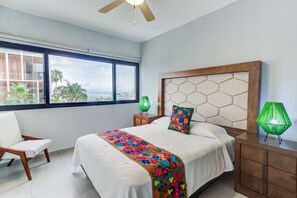 Relax in a serene coastal retreat with a large bed, wooden nightstand, and a view of palm trees and the ocean.