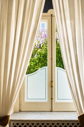 Blick durch ein Fenster mit Vorhängen, eine weiße Tür mit Messingklinke und ein Ausblick auf lila Blumen und Grünpflanzen draußen.