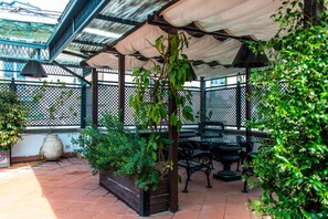 Ein überdachter Patio-Bereich mit Tisch und Stühlen, umgeben von Grünpflanzen und einem Metallzaun.
