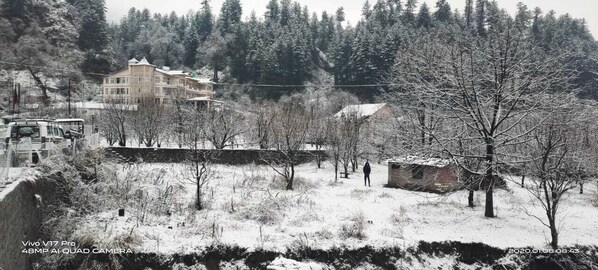 A snow-covered landscape with a building, trees, and a vehicle. - Snow Touch Resort By Sriniwasa Hotels (Kullu)