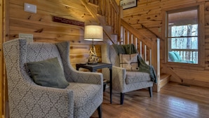 Een gezellig chaletinterieur met houten wanden, twee gestreepte fauteuils, een tafel met een lamp en een raam met uitzicht op bomen.