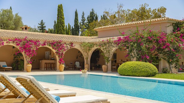 Un espace piscine avec des chaises longues, une cuisine et un patio couvert agrémenté d’arches et de plantes fleuries.