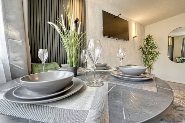 A modern dining area with a round table set for four, a potted plant, and a flat-screen TV mounted on the wall.