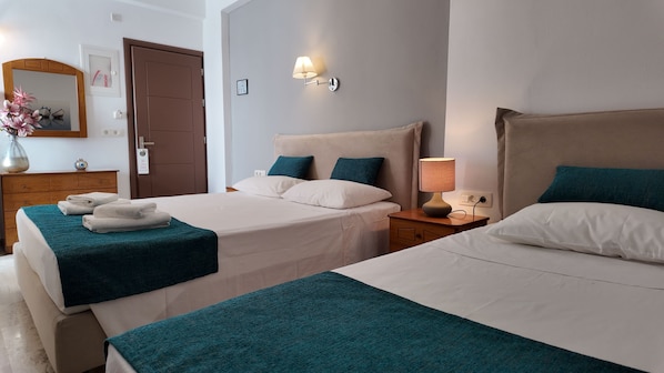 A hotel room with two beds, each with a teal bedspread and white pillows. There is a wooden dresser with a vase of flowers and folded towels on it. A wall-mounted lamp and a bedside table with a lamp are also visible.