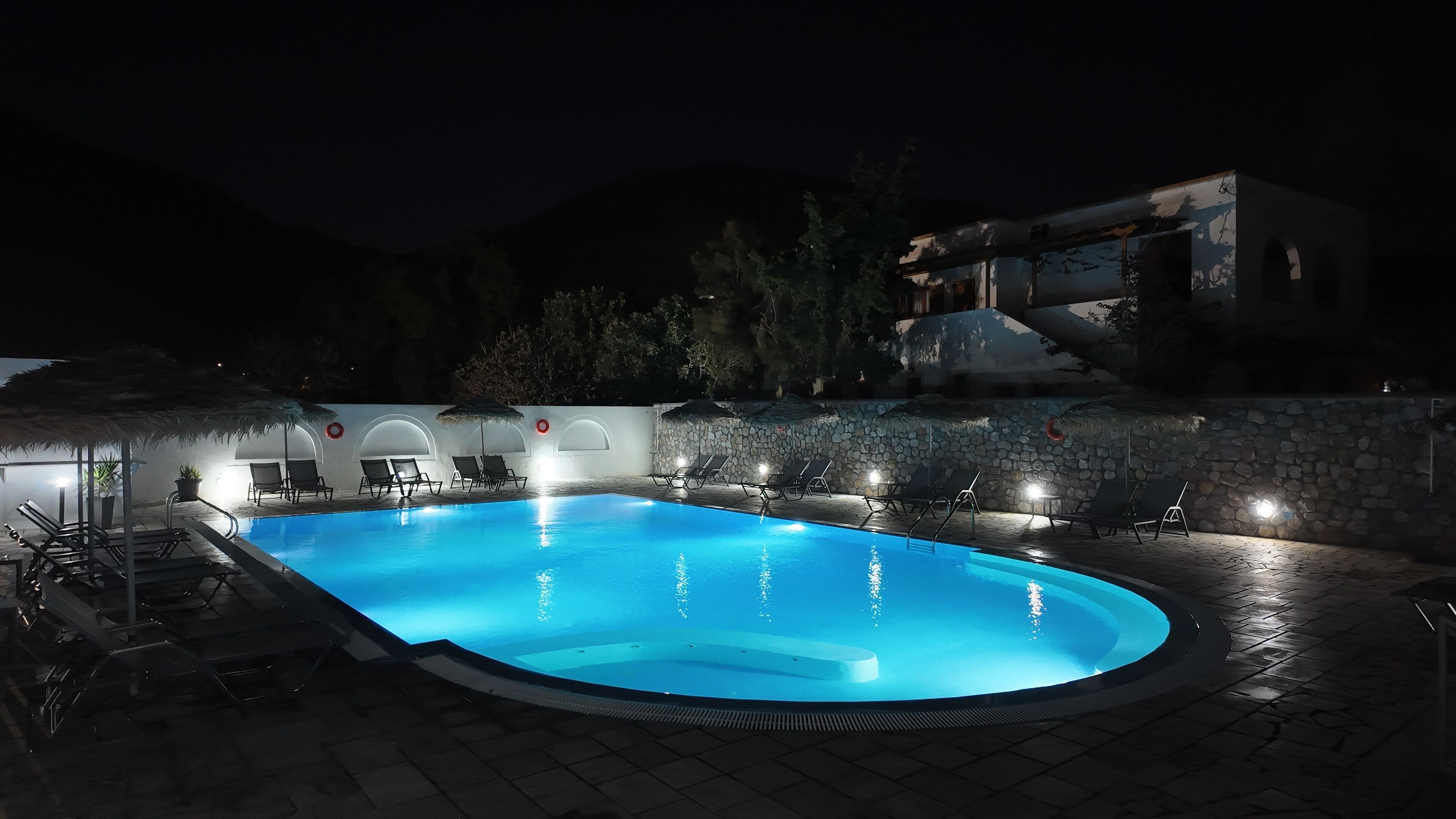 A nighttime swimming pool with lounge chairs and umbrellas, surrounded by stone walls and a mountain backdrop.
