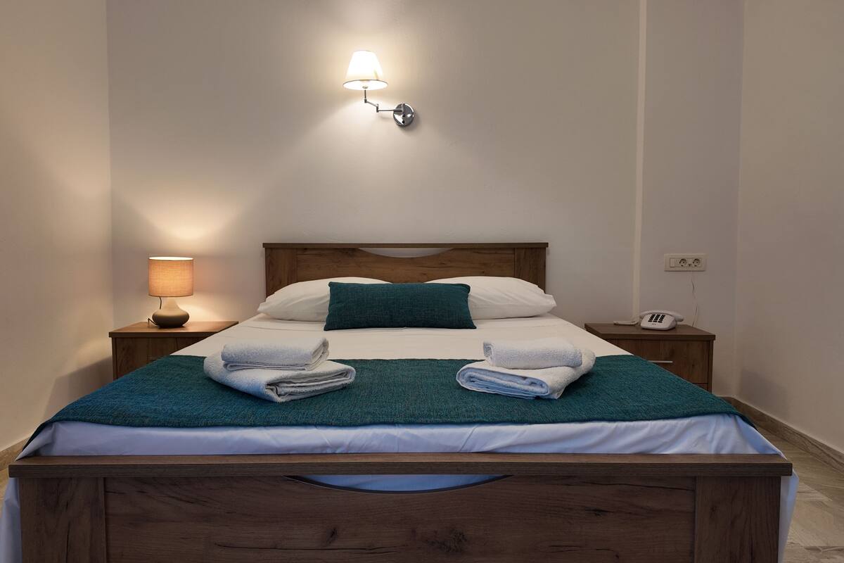 A neatly made bed with a wooden headboard, a teal throw blanket, and two folded white towels. There is a bedside lamp, a phone, and a wall-mounted light fixture.