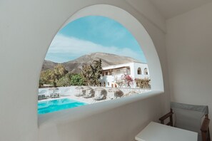 A view through a window of a white building with a pool and outdoor seating, overlooking a mountainous landscape.