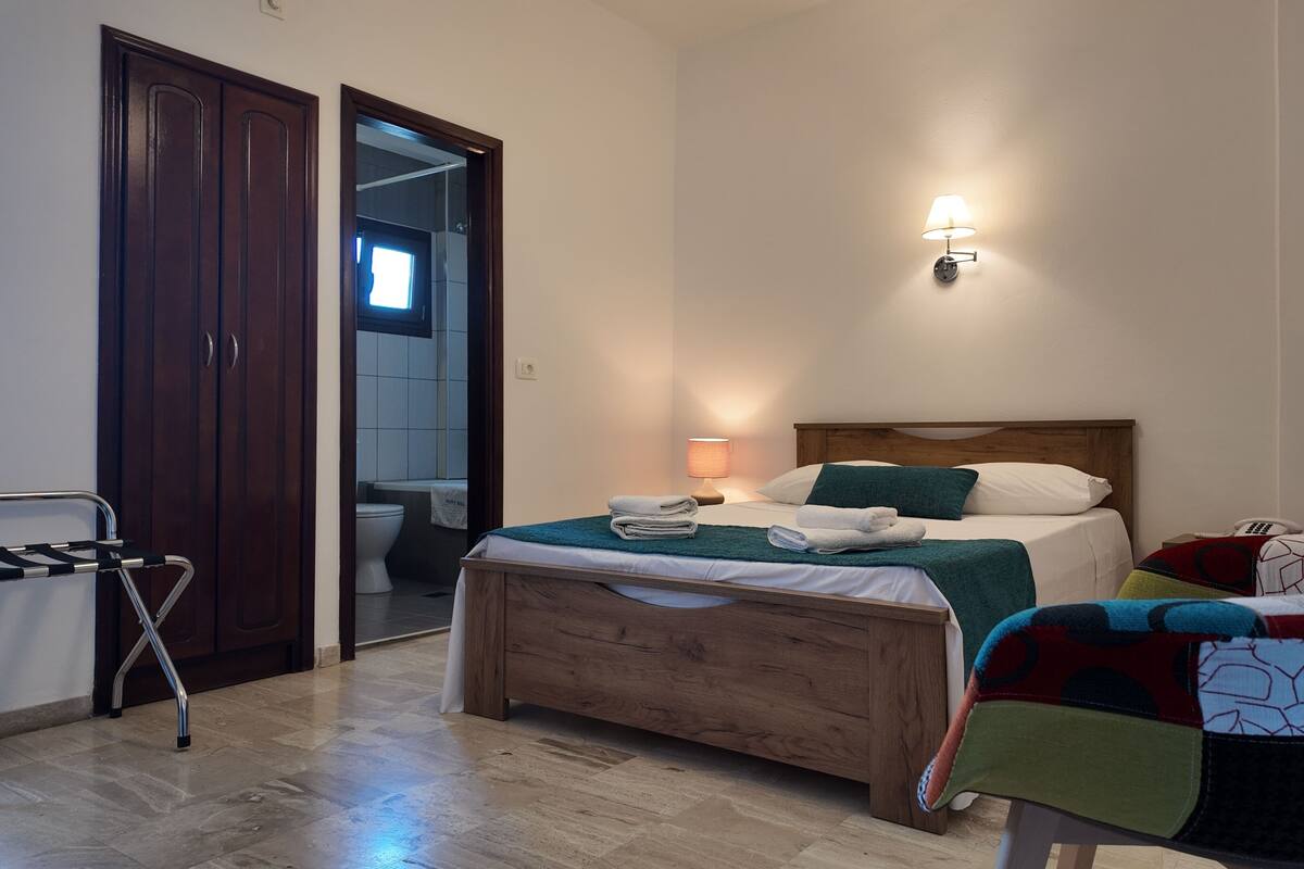 A bedroom with a wooden bed, a bedside table with a lamp, a chair with a colorful blanket, and a wooden wardrobe.