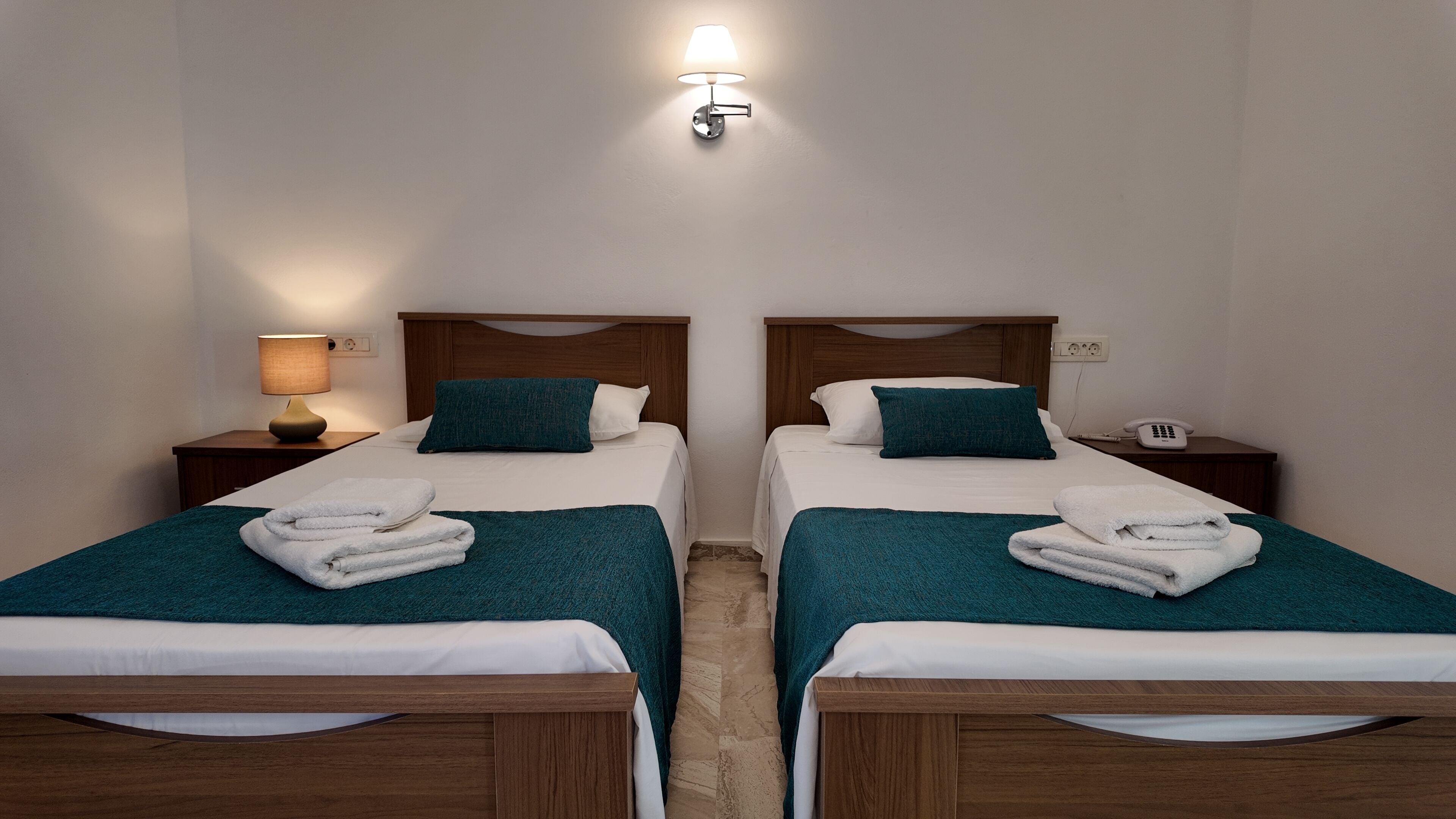 A hotel room with two single beds, each with a wooden headboard, white bedding, teal throw blankets, and neatly folded towels. There is a bedside lamp on the left side and a phone on the right side of the bed.