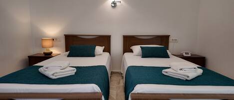 A hotel room with two single beds, each with a wooden headboard, white bedding, teal throw blankets, and neatly folded towels. There is a bedside lamp on the left side and a phone on the right side of the bed.
