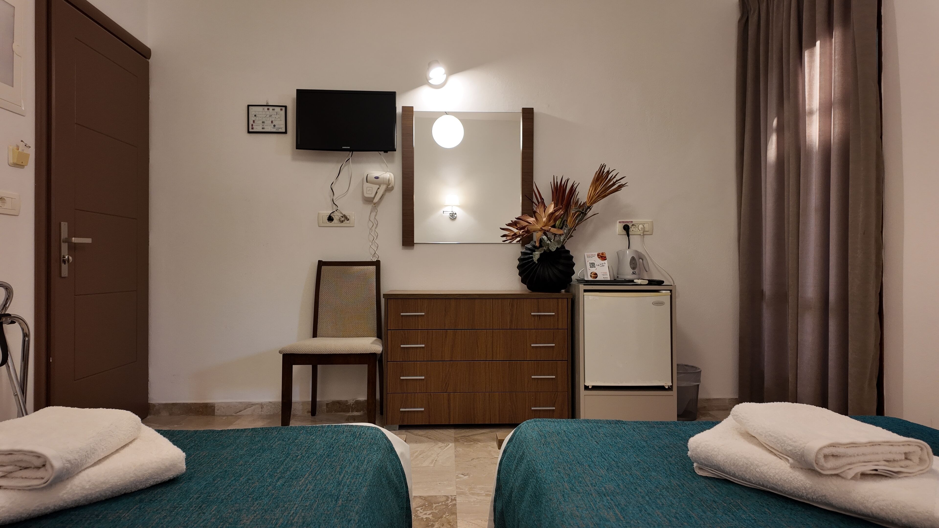 A hotel room with two beds, a TV, a mirror, a small refrigerator, a chair, and a potted plant.