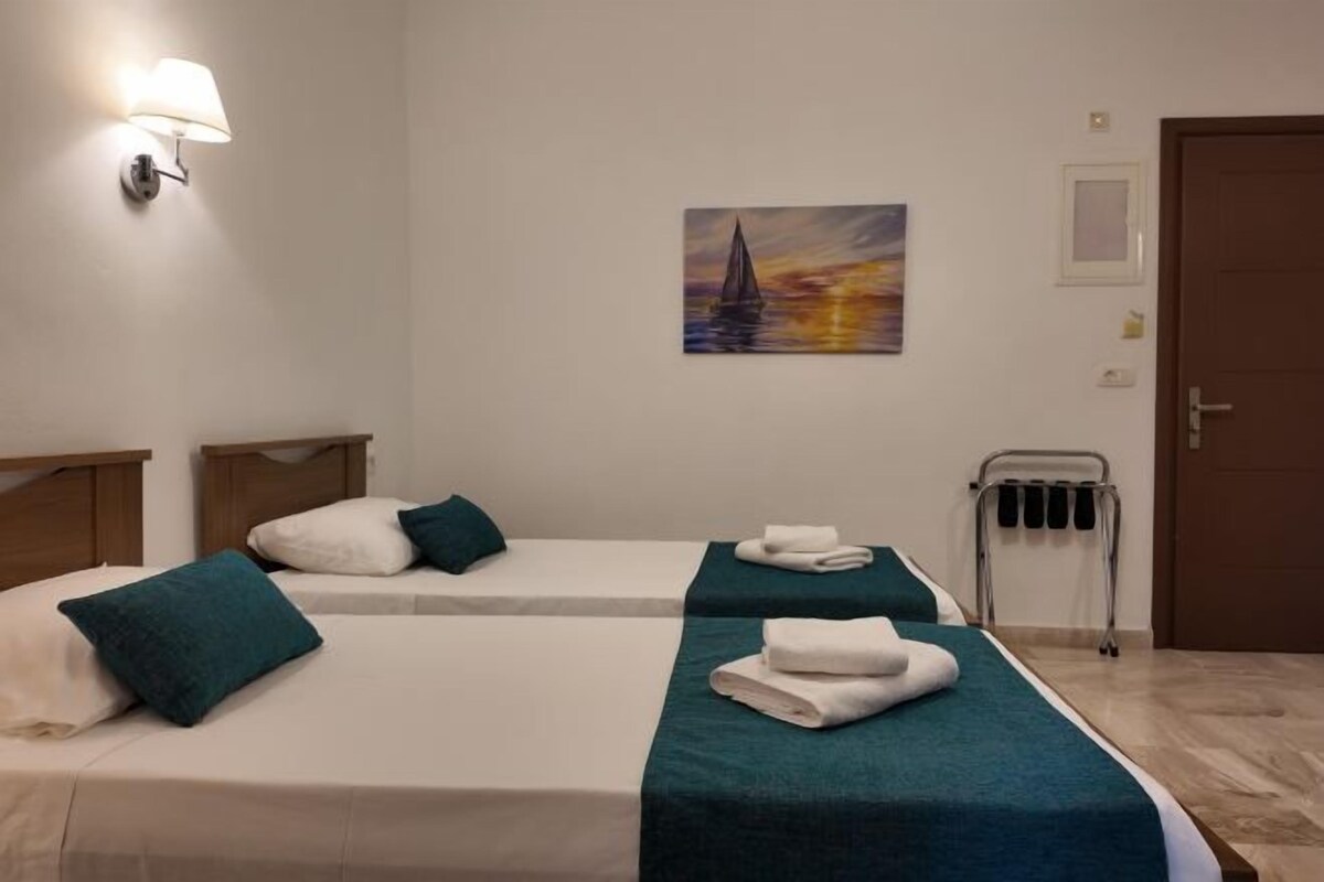 A hotel room with two beds, each with a teal bedspread and white linens, a painting of a sailboat on the wall, a wall-mounted lamp, and a door.