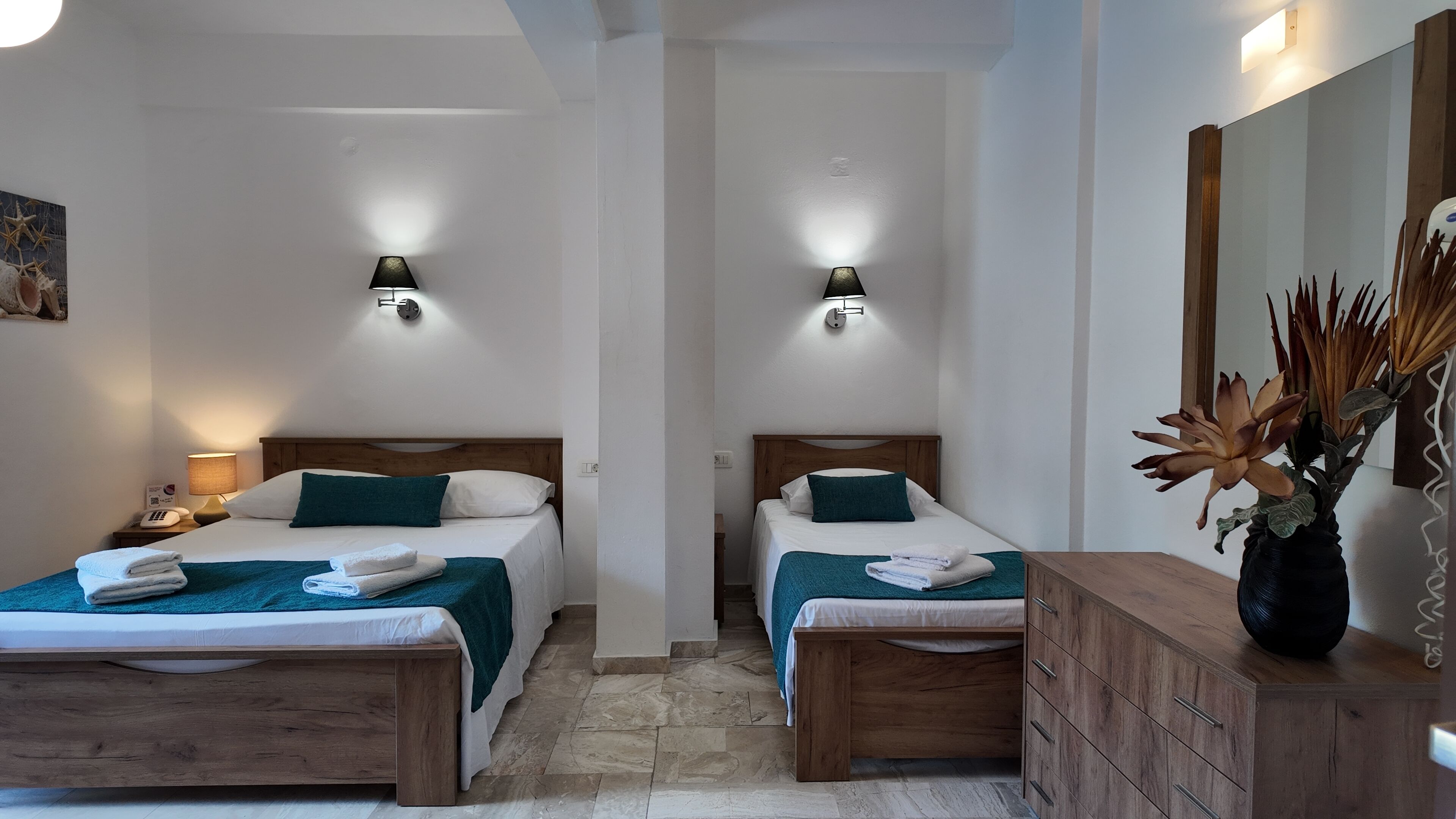 Ein Hotelzimmer mit zwei Betten, jeweils mit einem hölzernen Kopfteil, weißer Bettwäsche und Türkis-Kissen. Es gibt Nachttischlampen, einen Nachttisch aus Holz mit einem Telefon und eine Holzkommode mit einer Vase mit Trockenpflanzen.