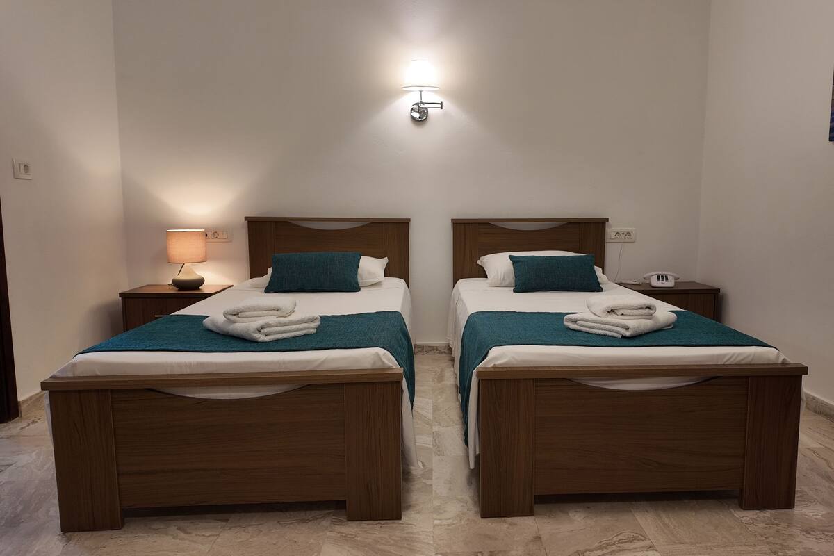 A hotel room with two wooden beds, each with a teal blanket and white towels. There is a nightstand with a lamp and a phone on the right side.