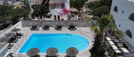 A rooftop swimming pool with lounge chairs and umbrellas, surrounded by white buildings and greenery.