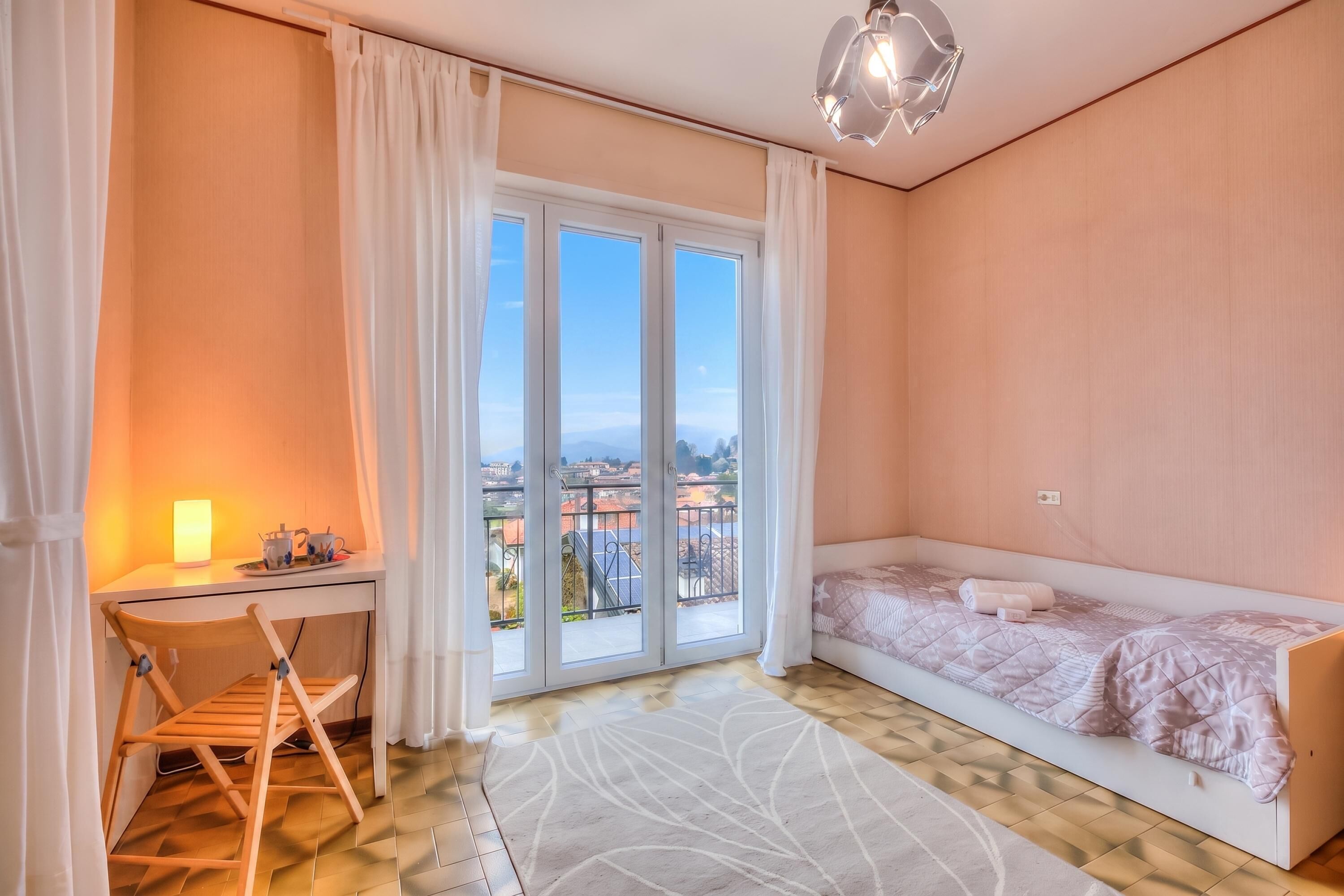 A bedroom with a bed, a desk, a chair, a window with a view, and a ceiling light.