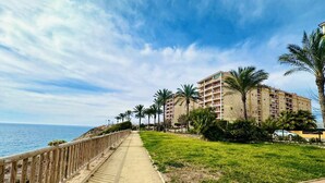 A coastal walkway with a wooden fence, a grassy area, and a multi-story building with balconies. - A1040 - First line of the sea in El Torres beach (Villajoyosa)
