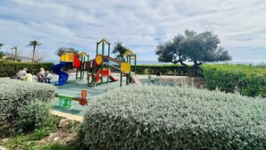 A playground with various play structures, surrounded by neatly trimmed hedges and a large tree. - A1040 - First line of the sea in El Torres beach (Villajoyosa)