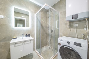 A modern bathroom with a glass-enclosed shower, a built-in washing machine, and a vanity with drawers. - Night Pearl - Deluxe Apartment (Bar)