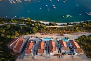Vista aérea de um hotel à beira-mar com vários edifícios, piscina e barcos na água.